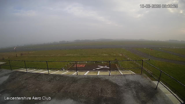 A view from a webcam at Leicestershire Aero Club showing a large grassy area of an airfield under a cloudy sky. In the foreground, there is a fenced area with a red and white marked section. A landing pad resembling a cross is visible in the center. The background features a wide, open expanse of land, with some blurred outlines of trees and a distant horizon. The overall scene has a grey and misty appearance, suggesting overcast weather.