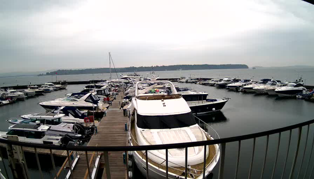 A marina filled with various boats and yachts docked along wooden piers. The scene is overcast, with a gray sky and calm water reflecting the surroundings. In the background, a distant landmass is faintly visible across the water's surface. There are several boats of different sizes, some with canopies and others without, creating a busy yet serene atmosphere.