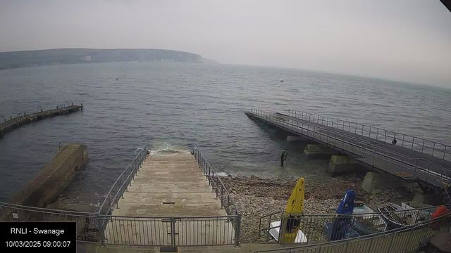 A coastal scene shows a grey, cloudy sky above the calm sea. In the foreground, there is a concrete ramp leading down to the water's edge, bordered by railings. To the right, a small yellow kayak is positioned against a rock-strewn beach. There is a person in dark clothing standing in the shallow water, near the edge of the ramp. In the background, a wooden pier stretches into the water, and a second, larger pier is visible to the left. A distant view of cliffs can be seen along the horizon.