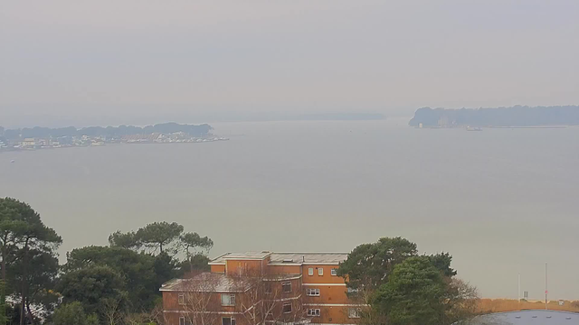 A hazy view of a body of water extending into the distance, with trees and buildings partially visible in the foreground. The shoreline on the left features a row of boats and structures, while a distant landmass can be seen on the right. The atmosphere appears overcast with muted colors, creating a soft, blurred appearance across the scene.