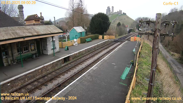 An image of Corfe Castle railway station on a cloudy morning. The view shows a quiet station platform with several green benches and a green sign reading "WAY OUT." In the background, there is a hill with the ruins of Corfe Castle at the top. A small blue building is visible on the left side of the platform, and a person dressed in orange can be seen near the railway tracks. The tracks run alongside the platform, leading off into the distance.