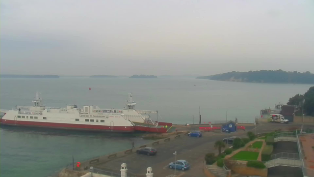 A view of a harbor with a ferry docked at the pier. The ferry is predominantly white with a red stripe and has a small control tower. In the background, there are some blurred green islands on the water horizon. The sky is overcast and gray, creating a muted atmosphere. In the foreground, there is a road with several cars parked, and a few pedestrians walking near the water. The pier has a small blue building and some red barriers along the edge. There is greenery and landscaped areas visible on the right side of the image.