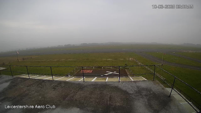 A view from a webcam showing a grassy airfield under overcast skies. In the foreground, there is a railing and a helipad marked with a white cross. The background features a flat expanse of grass and a distant, hazy view of more grass and a few defined areas of darker surfaces, likely runway or taxiway. Visibility is limited due to fog. The bottom left corner has text indicating "Leicestershire Aero Club" along with the date and time.