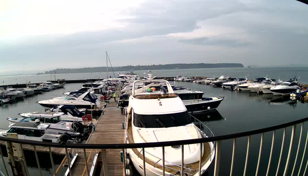 A view of a marina with several boats docked in calm water. The foreground features a white yacht with a tan-colored top, and the wooden pier extends to the left. Many other boats, both large and small, are scattered throughout the marina. The sky is overcast, and there are distant hills visible in the background.