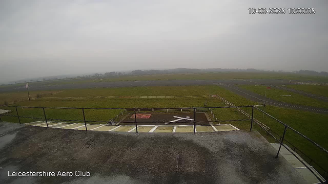 A view from a webcam shows a large grassy field with a few patches of gravel and a tarmac area in the foreground. In the distance, there is a flat landscape with a low horizon, obscured by a grey sky that indicates overcast weather. A white "X" marking is visible on the ground, surrounded by a wooden fence. On the left, there is a railing of a raised platform, and in the background, thin white lines correspond to runways or roads, while a small flag can be seen flapping in the breeze. The overall atmosphere is calm and tranquil, with a muted color palette.