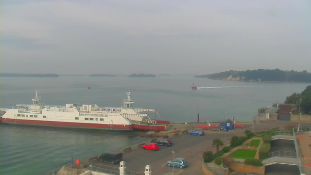 A view of a coastal area featuring a white ferry with red accents docked at a harbor. The water is calm and reflects the light, with some small islands visible in the background. A smaller boat is seen moving across the water, leaving a white wake behind it. In the foreground, a parking area is filled with cars of various colors, including red and blue, while greenery and pathways are visible near the water's edge. The sky is overcast, creating a muted atmosphere.