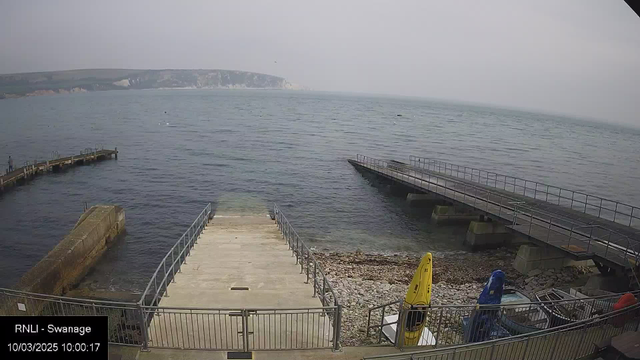 A coastal scene with calm waters, showing a concrete ramp leading down to the shoreline. To the left, there's a small wooden pier extending into the water, while another pier is visible to the right. A rocky beach is in the foreground, with several colorful kayaks in yellow and blue parked on the side, next to a boat. The backdrop features a gently sloping green hillside and a cliff in the distance, beneath a muted gray sky.