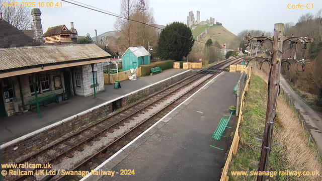 A railway station platform featuring a stone building with a sloped roof on the left. There are green benches along the platform, and a green shed in the background. The tracks run parallel to the platform, with gravel and wooden sleepers visible. On the hillside in the distance, there are ruins of a castle. The sky is clear with a few clouds, and the overall scene appears peaceful and rural.