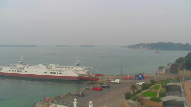 A ferry is docked by a harbor, with a few vehicles parked nearby. The water is calm and stretches out towards the horizon, where small islands can be seen in the distance. The sky appears hazy. In the foreground, there are green plants and pathways leading around the area. Several other boats are visible further out on the water.