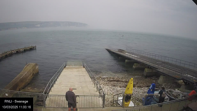 A view of a seafront area with calm waters. In the foreground, there is a concrete ramp leading down to the water, bordered by a metal railing. To the right, there are bright yellow and blue kayaks resting on the shore. A person stands on the ramp, facing the water. In the background, a long wooden pier extends into the sea, and there are faint hills visible in the distance, partially obscured by misty weather. The overall atmosphere is quiet and serene.