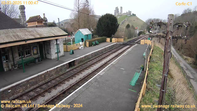 A railway station platform with green benches and a stone building on the left. There is a wooden shed painted blue in the background. In the distance, a hill with ruins of a castle is visible, surrounded by trees. The track runs along the platform with wooden fencing nearby. The sky is overcast, and a telephone pole is positioned on the right side of the image.