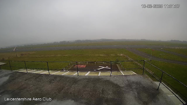 A cloudy and overcast view from a webcam positioned at an elevated angle, overlooking a grassy airfield. In the foreground, there is a railing and a marked area with a white cross symbol on a darker surface. Beyond that, the airfield is visible, featuring a mixture of grass and asphalt runways. The background is hazy, blending with the grey sky, suggesting poor visibility. The presence of some scattered objects and a few signs can be identified along the edges of the airfield.