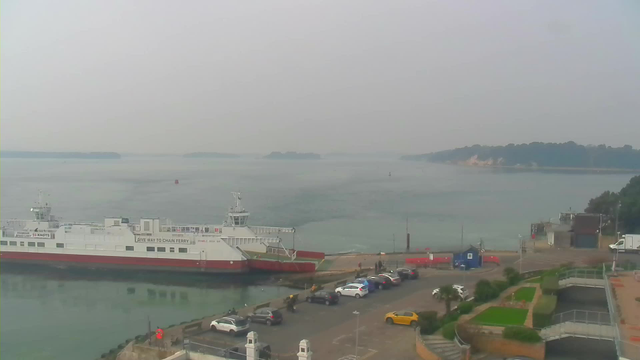 A view of a harbor with a large ferry docked at the left side, featuring a red and white design. The water is calm and appears slightly murky, reflecting overcast skies. In the foreground, there are several parked cars along a waterfront road, while a few people walk in the area. The background shows a distant shoreline with trees and beige cliffs. The image has a hazy atmosphere, suggesting limited visibility due to fog or mist.