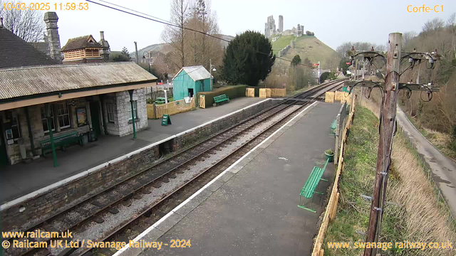 A view of a train station with a stone platform and several green benches. On the left, there is a building with a sloped roof, and further back, a turquoise shed. A wooden fence surrounds a small area, and a sign that reads "WAY OUT" is visible. The tracks run parallel to the platform, and in the background, the remains of a castle can be seen on a hill. The scene is set on a clear day with trees in the vicinity.