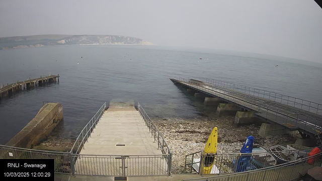 A view of a calm sea with a foggy sky, featuring a concrete boat launch area with a railing. To the right, a small wooden pier extends into the water. There are several boats moored in the distance. The shoreline is rocky, with visible pebbles and a couple of bright kayaks on display: one yellow and one blue. The date and time are displayed in the lower left corner.