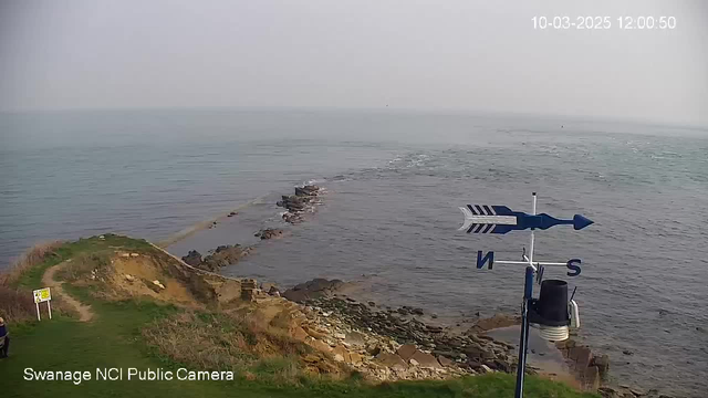 A coastal landscape showing a rocky shoreline extending into the sea. The water appears calm with gentle waves. In the foreground, there is a grassy area leading to the edge of a cliff, with a white signpost visible. To the right, a weather vane with arrows indicating wind direction stands. The sky is hazy, and the scene seems tranquil and natural. The date and time are displayed in the top right corner.