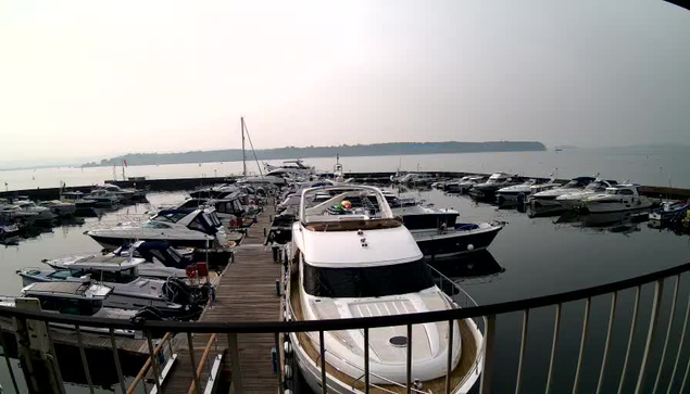 A panoramic view of a marina with numerous boats docked. In the foreground, there are several white and dark-colored yachts moored at a wooden pier. The water is calm, reflecting the boats and a light gray sky, indicating a hazy atmosphere. Further in the background, land can be seen across the water, and a few masts and flags are visible among the boats. The scene captures a quiet, overcast day at the marina.