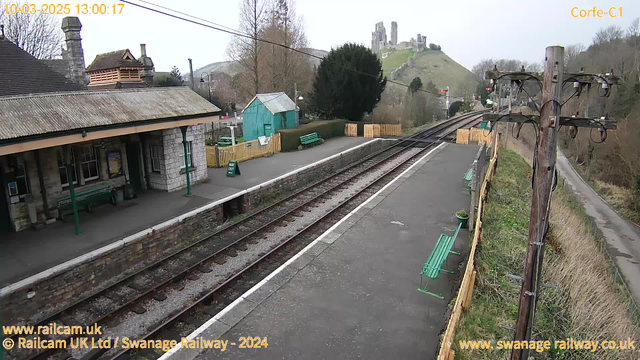 A view of a train station with a platform featuring a stone building on the left. There are green benches along the platform and a wooden fence in the background. A small turquoise shed is positioned nearby, and a sign reads "WAY OUT." In the distance, there is a hill with ruins of a castle or fort on top. The scene is set under an overcast sky.