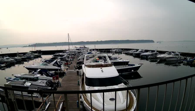 A marina scene featuring numerous boats docked in calm waters. In the foreground, several white and blue boats are visible, including a large white yacht. The background shows a misty horizon and a hazy sky, with a few distant hills and boats further out on the water. The marina is lined with wooden docks.