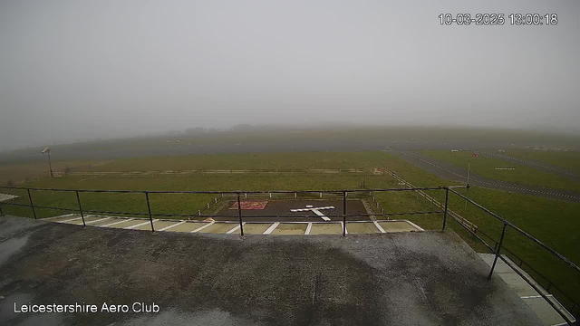 A foggy view from a webcam at the Leicestershire Aero Club. The foreground features a flat, concrete area with a white cross marked on the ground. There is a railing in front, and beyond it, a grassy field extends into the fog, making it difficult to see far. A wind sock is visible on the left side, indicating light wind. The background is dominated by thick fog, obscuring most details and creating a gloomy atmosphere.