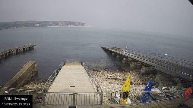 A coastal scene at Swanage shows a calm sea under a hazy sky. In the foreground, a concrete pathway leads down to the water, flanked by a railing. To the left, a wooden pier extends into the sea, with some boats visible near the shore. On the right, two colorful kayaks, one yellow and one blue, are resting on the beach. The shoreline is rocky, and the distant cliffs appear blurred due to mist.