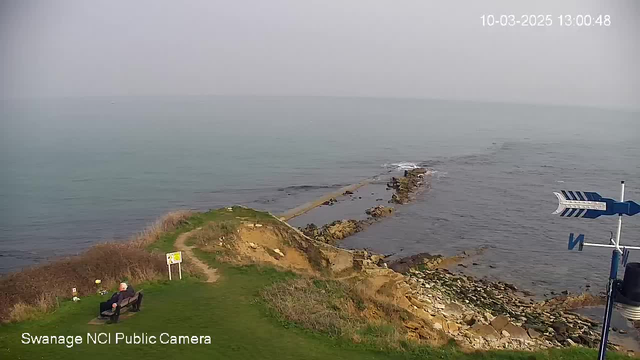 A serene coastal scene captured from a webcam, showing a grassy area with a bench facing the sea. To the left, there is a pathway leading towards rocky outcrops extending into the water. Gentle waves can be seen lapping against the rocks, and the horizon blends into a hazy sky. A small sign is positioned near the bench, and in the foreground, there are some flowers. In the lower right corner, a weather vane indicates North with blue and white markings.