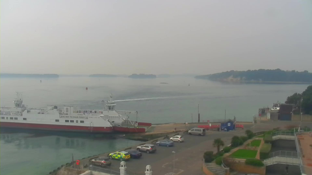 A view of a calm harbor featuring a large white and red ferry docked near the shore. In the background, there are several small boats and buoys on the water under a hazy sky. Parts of a coastal area, including trees and structures, are visible along the shoreline. A small parking lot with several cars, including a police vehicle, is in the foreground. The scene conveys a tranquil atmosphere with minimal waves on the water.