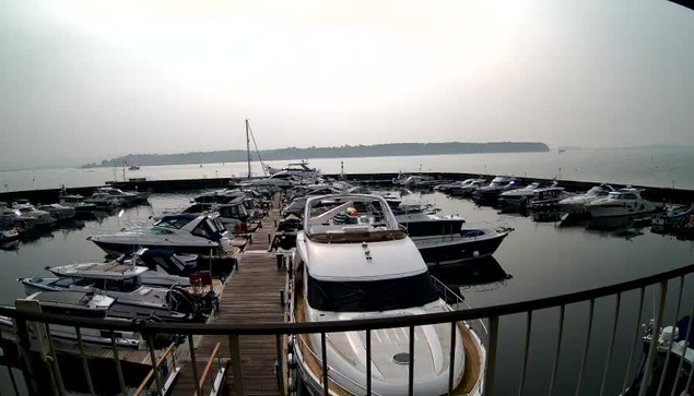 A view of a marina filled with various boats and yachts docked at a wooden pier. The water is calm and reflects the boats. The background features a hazy sky, suggesting overcast weather, with land visible in the distance. The scene conveys a peaceful, nautical atmosphere.