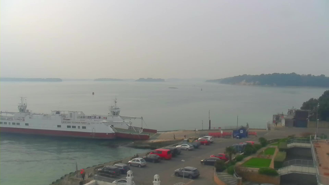 A serene waterfront scene shows a large white ferry with red accents docked at a pier. The calm water extends into the distance, reflecting the grayish sky. In the foreground, there is a parking lot filled with various cars, mostly in shades of gray and red. To the right, a small structure and some greenery contribute to the coastal setting. In the background, gentle hills and trees are visible along the shoreline. The atmosphere is hazy, suggesting overcast weather.