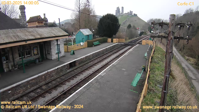 A train station scene with a stone platform featuring green benches. There is a vintage-style building on the left, and a blue shed in the background. Several empty railway tracks extend into the distance. A sign stating "WAY OUT" is visible, and a distant hill with castle ruins is present in the background. The sky appears overcast.