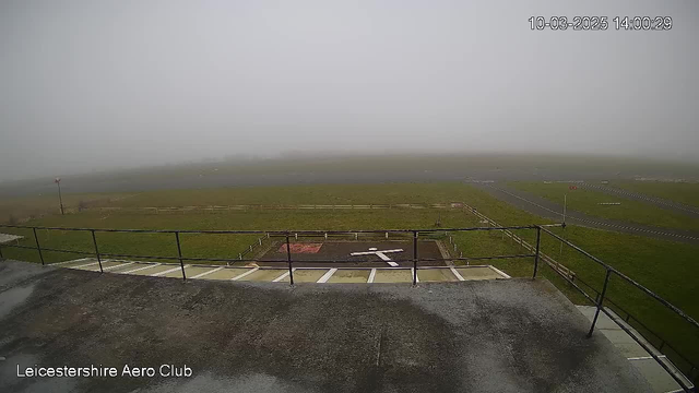 A foggy view of an aerodrome, showing a grassy runway and a helipad marked with a white cross. The background is obscured by dense fog, making distant features indistinct. A fence lines the perimeter of the aerodrome, with a few signs visible. The scene is gray and muted due to the fog, creating a subdued atmosphere.