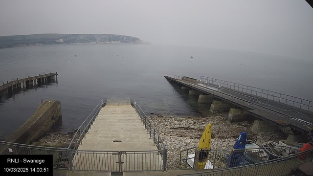 A view of a calm coastline with a light gray sky. In the foreground, there is a wide concrete ramp leading down to the water, flanked by metal railings. To the left, there is a stone pier extending into the water, while to the right, a wooden platform with a railing also extends over the water. Various small boats are visible in the water, and colorful kayaks—one yellow and one blue—are positioned on the shore next to the ramp. The rocky shoreline is visible, and a distant cliffside can be seen in the background.