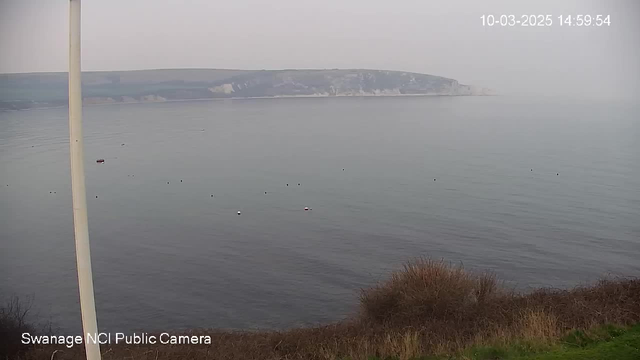 A hazy view of a calm sea with a distant shoreline visible. The water appears smooth and mostly still, with a few small boats scattered throughout. In the foreground, there are some brownish grass and shrubs. The scene is muted due to fog or mist, giving it a serene but overcast atmosphere. A tall white pole is present on the left side of the image. The date and time are displayed in the upper right corner.