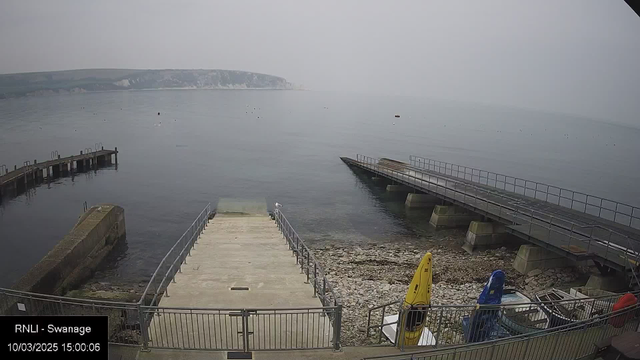 A coastal scene shows a quiet, misty harbor with calm waters. In the foreground, there are stone steps leading down to the water, surrounded by a metal railing. To the right, two kayaks are visible, one yellow and one blue, resting on the shore. In the background, multiple piers extend into the water, with a rocky shoreline and a grassy cliff rising in the distance. The sky is hazy, and small boats are visible on the water's surface. The atmosphere appears peaceful and serene.