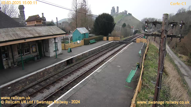 A view of a train station platform with several green benches along the edges. There is a stone building with a sloped roof on the left, and a small wooden shed painted teal behind it. A wooden fence surrounds part of the area, with a sign that reads "WAY OUT" visible. In the background, there are hills with ruins at the top, partially obscured by trees. The skies are cloudy, and railway tracks run parallel to the platform, leading into the distance.