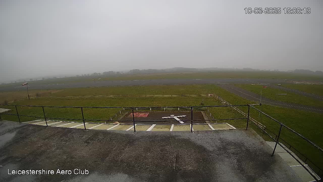 A view from a webcam at the Leicestershire Aero Club shows a foggy landscape. In the foreground, there is a fenced-off area with a white cross shape on the ground, likely a landing zone or helipad. The grass surrounding the area is green, and a small portion of a paved runway is visible in the distance. There are limited visibility conditions due to fog, obscuring any features beyond the immediate vicinity. A flagpole with a flag is also seen to the left of the image.