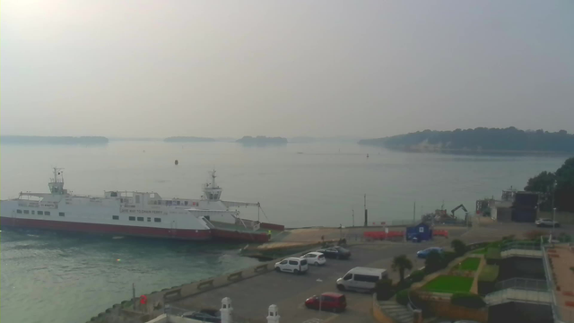 A view of a calm body of water with a hazy sky. In the foreground, a white ferry with a red stripe is docked at a pier. Several vehicles are parked nearby, including two white cars and one blue car. To the right, there is a construction area with machinery visible. In the background, distant islands can be seen through the mist, and the water reflects the muted light of the overcast sky.