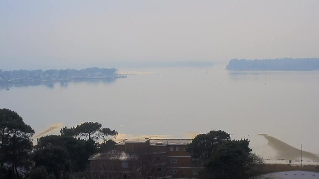 A tranquil seascape viewed from a height, with a hazy, foggy atmosphere. In the foreground, there are dark green trees and a low building with windows. The middle ground features a calm water body reflecting the muted grey sky, with hints of soft yellow sandbanks emerging at low tide. In the distance, a shoreline with scattered houses and boats is visible, along with an island partially obscured by fog. The overall mood is serene and quiet, with no visible people or animals.