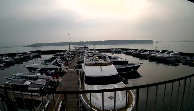 A view of a marina filled with various boats and yachts docked along wooden piers. The water is calm and reflects the overcast sky above, which appears hazy. In the background, green hills are visible along the horizon, while a few boats are moored further out. There is a sense of tranquility in the scene, with soft lighting and a muted color palette.