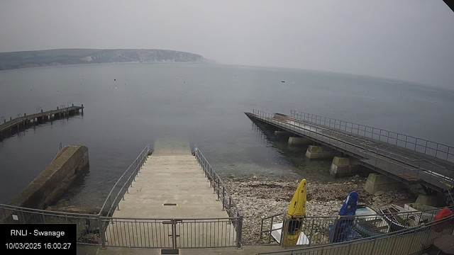 A view of a calm body of water with a rocky shore in the foreground. There are two wooden docks extending into the water, one on the left and another on the right. A concrete ramp leads down to the water from the shore. Several kayaks are stacked on the side, with one yellow kayak visible. The sky is overcast, contributing to a muted atmosphere, and the distant land features cliffs, partially obscured by fog.