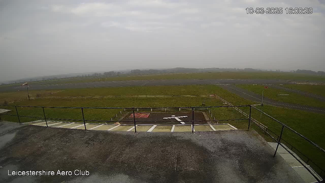 A view from a webcam overlooking an airfield. The foreground features a railing around a concrete area with a large white cross marked on the ground. The background shows a grassy area with some scattered trees and a cloudy sky, creating a somewhat overcast atmosphere. The airfield has a runway and taxiway that are mostly visible, with a few parked vehicles in the distance.