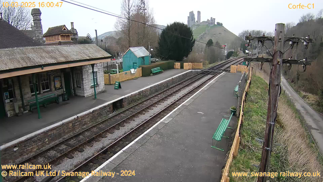 A railway station platform with a stone building featuring a sloped roof on the left side. Green benches are placed along the platform, and a sign indicating "WAY OUT" is visible. In the background, a hill rises featuring a castle ruin at its peak. The landscape includes trees and a blue shed. There are railway tracks running parallel to the platform, with the scene set on a cloudy day. The time and date in the corner indicate it is March 10, 2025, at 4:00 PM.