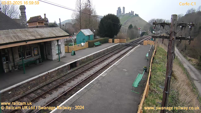 A view of Corfe Castle railway station, featuring a stone platform with green benches and a roof. The tracks run through the middle of the image, leading away from the station. In the background, Corfe Castle sits atop a hill with greenery around it. There are trees and bushes lining the platform, and a wooden fence separates the area from a small road. A sign that reads "WAY OUT" is visible on the platform. The scene is set on a cloudy day.