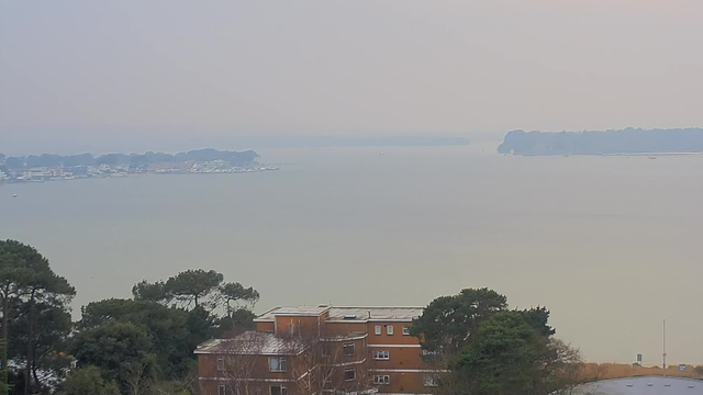 A view of a calm body of water extending towards the horizon, with a hazy sky above. In the foreground, there are trees and a brown building with multiple stories. To the left, there is a shoreline with several boats and a distant landmass outline, possibly an island, visible in the background. The overall atmosphere is tranquil and somewhat overcast.