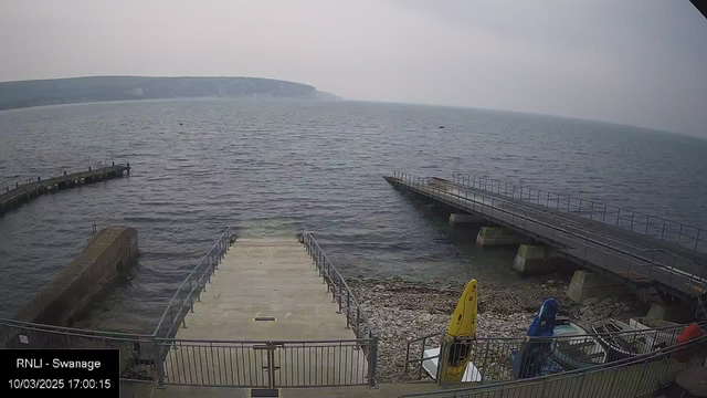 A coastal view featuring a calm sea under a cloudy sky. A wide stone jetty extends into the water from the left side. On the right, there is a wooden pier with a railing. Two colorful kayaks, one yellow and one blue, are stored beside the jetty. The shore is rocky, with pebbles visible. The landscape in the background consists of low cliffs and hills, partially obscured by mist. The scene is tranquil and lacks any visible people or boats.