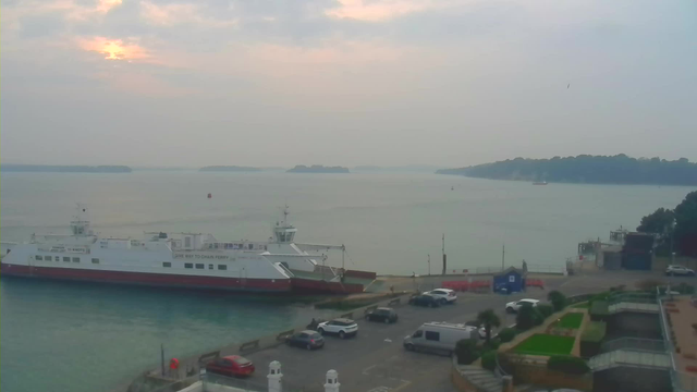 A large ferry boat is docked at a port, with its white body and red bottom clearly visible. The calm water reflects a cloudy sky with soft sunlight peeking through. In the background, there are distant islands and a hazy shoreline. Near the ferry, several parked cars are visible on the pavement, and there are some plant beds and steps leading down to the water. The scene conveys a peaceful waterfront atmosphere.