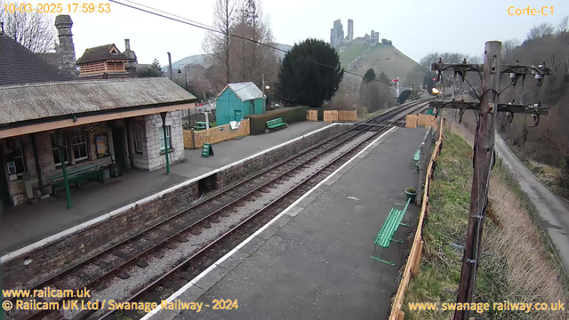 A view of a train station with a stone platform and wooden benches painted green. To the left, there is a building with a pitched roof and a chimney, featuring windows and a bench outside. There are also green benches along the platform. In the background, a hillside with old stone ruins can be seen, along with trees and a wooden fence. On the platform, a "WAY OUT" sign is visible, and there are train tracks leading off into the distance. The scene is set in a rural area, with a gray sky above.