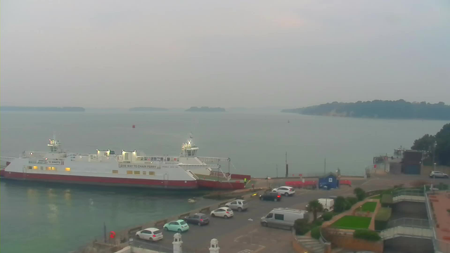 A waterfront scene showing a chain ferry docked at a harbor. The ferry has a white deck with red trim and several lights illuminated. In the background, calm waters extend to distant, hazy islands. There are parked cars along the harbor, and greenery is visible in the foreground, with some steps leading down from a raised area. The sky is overcast, creating a muted atmosphere over the landscape.