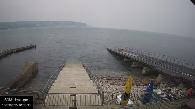 A view of a harbor scene with calm water. In the foreground, a set of concrete steps leads down to the water, flanked by railings. To the left, a curved dock extends into the water, with several posts visible. On the right, a straight wooden pier is positioned over the water, with railings along the edge. There are various boats, including a yellow kayak and a blue kayak, stored along the shoreline. The sky appears overcast, creating a muted atmosphere, and a distant shoreline can be seen in the background.