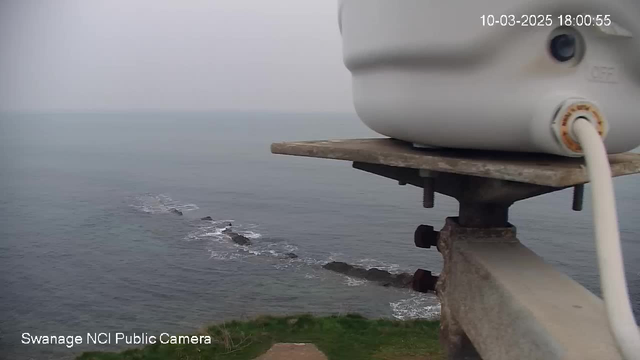 A close-up view of a white cylindrical object mounted on a metal platform, with a cable extending from it. In the background, there is a gray sky and a calm sea, where dark rocks are visible along the water's edge. The scene is slightly hazy, suggesting a quiet, overcast day. The bottom left corner contains the text "Swanage NCI Public Camera," and there is a timestamp in the top right corner indicating the date and time.